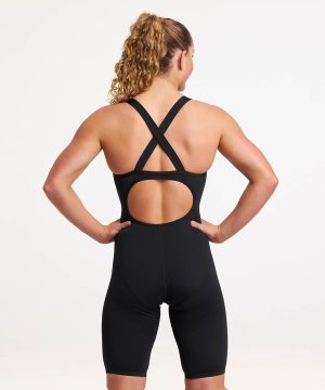 En kvinna med lockigt blont hår uppsatt i hästsvans står med händerna på höfterna och visar den öppna ryggen och de korsade banden på Funkita Still Black Legsuit för dam. Hon har denna svarta, ärmlösa sportiga dräkt mot en enfärgad vit bakgrund.